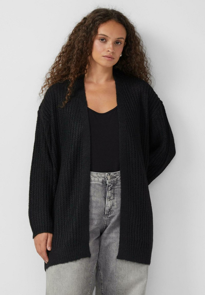 Czarny sweter z dzianiny z otwartym przodem, z oversize'owymi rękawami i teksturowanym wzorem. Noszony na czarnej bluzce i szarych dżinsach o wysokim stan.