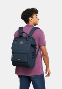 Navyblauer Rucksack mit strukturiertem Design, vorderer Reißverschlusstasche und Lederapplikationen; ausgestattet mit verstellbaren Trägern und einem oberen Griff.