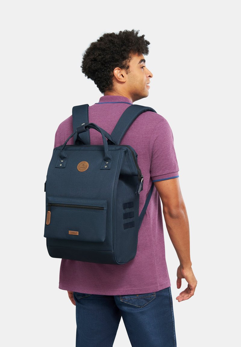 Navyblauer Rucksack mit strukturiertem Design, vorderer Reißverschlusstasche und Lederapplikationen; ausgestattet mit verstellbaren Trägern und einem oberen Griff.