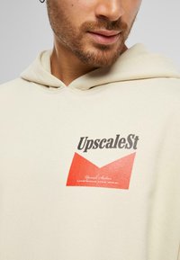 Beige hettegenser med frontlogo som viser "UpscaleSt" i svart og en rød trekantform. Mykt stoff med avslappet passform og fastsatt hette.