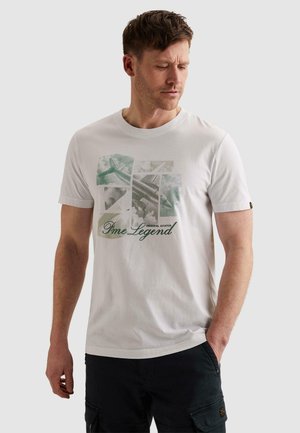 Man draagt een wit T-shirt met groene vliegtuigcollage en de tekst "Pme Legend", gecombineerd met zwarte cargobroek, staand met één hand in de zak.