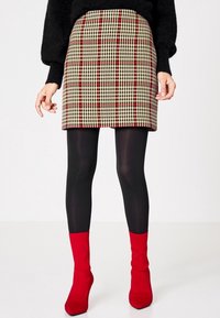 Femme portant une jupe à carreaux beige et rouge, des collants noirs, un pull noir et des bottes à talons rouges vives, se tenant contre un fond blanc.