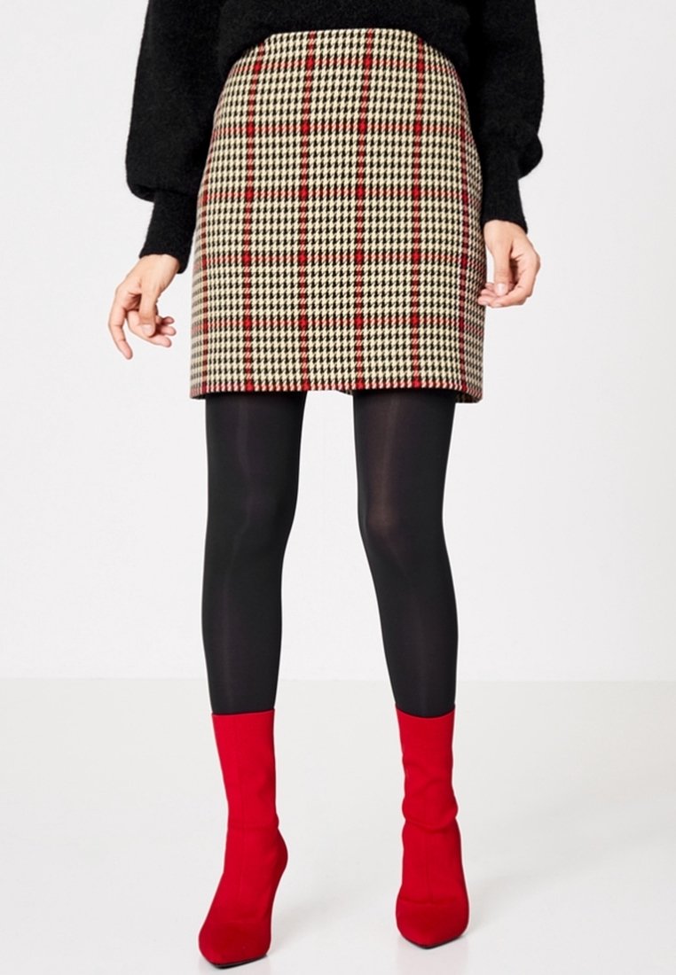 Femme portant une jupe à carreaux beige et rouge, des collants noirs, un pull noir et des bottes à talons rouges vives, se tenant contre un fond blanc.