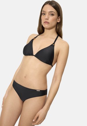 Mujer con cabello largo y castaño, vistiendo un bikini negro de triángulo y braguitas a juego, sobre un fondo blanco liso.