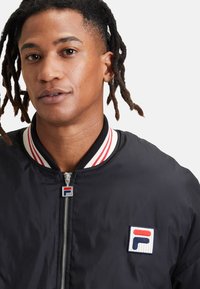Hombre con rastas que lleva una chaqueta negra de FILA con cuello a rayas rojas y blancas, y el logo de FILA en la cremallera y el pecho.