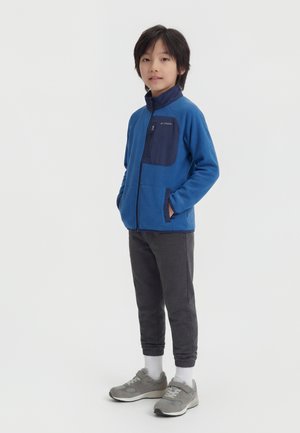 Poika seisoo ja katsoo kameraan sininen vetoketjullinen takki päällään, tummat verkkarit, valkoiset sukat ja harmaat lenkkarit yksiväristä taustaa vasten.