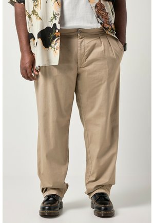 Osoba ubrana w beżowe plisowane spodnie, czarne skórzane buty sznurowane, białą prążkowaną koszulkę oraz wzorzystą koszulę zapinaną na guziki z nadrukiem tygrysa.