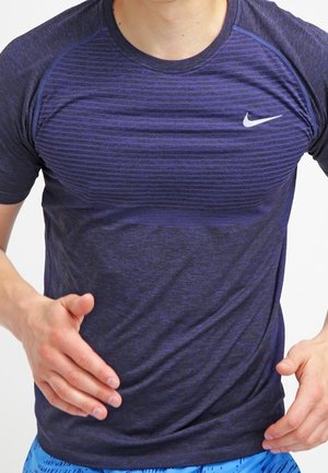 Mężczyzna biegnący, ubrany w ciemnoniebieską sportową koszulkę Nike z krótkim rękawem i niebieskie szorty z wzorem, widoczna górna część ciała, białe tło.