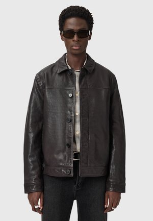 Dark brown leather jacket with a collared neck, front button closure, and two side pockets. Worn over a beige patterned shirt.