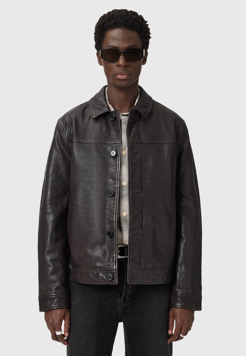 Veste en cuir marron foncé avec un col, fermeture à boutons à l'avant et deux poches latérales. Portée sur une chemise beige à motifs.