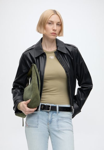 Femme blonde portant une veste en cuir noire, une chemise vert olive, un jean bleu clair, et tenant un sac à bandoulière vert olive sur un fond blanc.