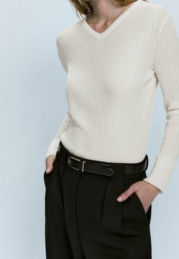 Personne portant un pull crème côtelé à manches longues avec col en V, rentré dans un pantalon noir taille haute avec une ceinture noire, mains dans les poches.