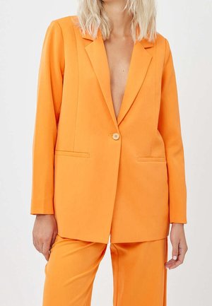 Blazer arancione con revers a punta, chiusura con un bottone e due tasche frontali. Tessuto liscio, vestibilità su misura, abbinato a pantaloni coordinati.