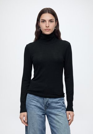 Femme aux longs cheveux foncés portant des boucles d'oreilles argentées, un pull à col roulé noir et un jean bleu, debout devant un fond blanc uni.