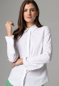 Weißes Hemd mit spitzem Kragen, Rüsche an den Ärmelabschlüssen und Knopfverschluss. Glattes, texturiertes Material. Modell posiert mit erhobener Hand.