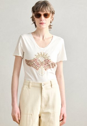 T-shirt bianca con scollo a V, decorata con ricami dettagliati di un sole e ali in tonalità rosa e oro, abbinata a pantaloni beige chiaro.