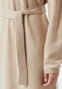 Beżowa dzianinowa sukienka z długimi rękawami i dopasowanym paskiem do wiązania. Charakteryzuje się miękką teksturą i prążkowanymi mankietami, podkreślającymi minimalistyczny design.