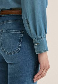 Jeansskjorte i lyseblå med lange, samlede ærmer og knapdetaljer ved manchetterne, parret med mørkeblå jeans med en baglomme.