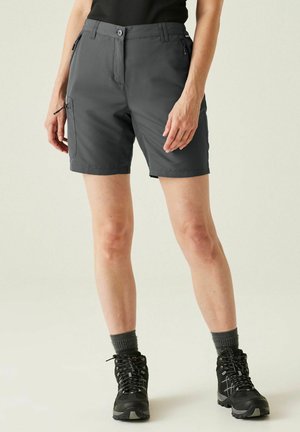 Persona que lleva pantalones cortos tipo cargo con cremallera de color gris oscuro, calcetines gris oscuro y botas de senderismo negras, de pie frente a un fondo liso.