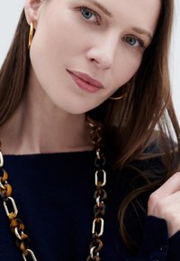 Modell mit langen braunen Haaren, die einen marineblauen Pullover trägt. Sie hat eine klobige Schildpatt-Halskette und goldene Creolen im Ohr und präsentiert einen neutralen Make-up-Look.