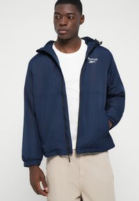 Tânăr îmbrăcat cu o jachetă Reebok bleumarin cu fermoar peste un tricou alb și pantaloni bej, având o mână în buzunarul jachetei.
