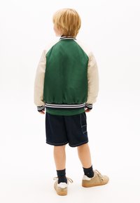 Kind mit kurzen blonden Haaren, trägt eine grün-cremefarbene Jacke, dunkle Shorts, schwarze Socken und beigefarbene Turnschuhe, steht mit dem Rücken zur Kamera vor weißem Hintergrund.