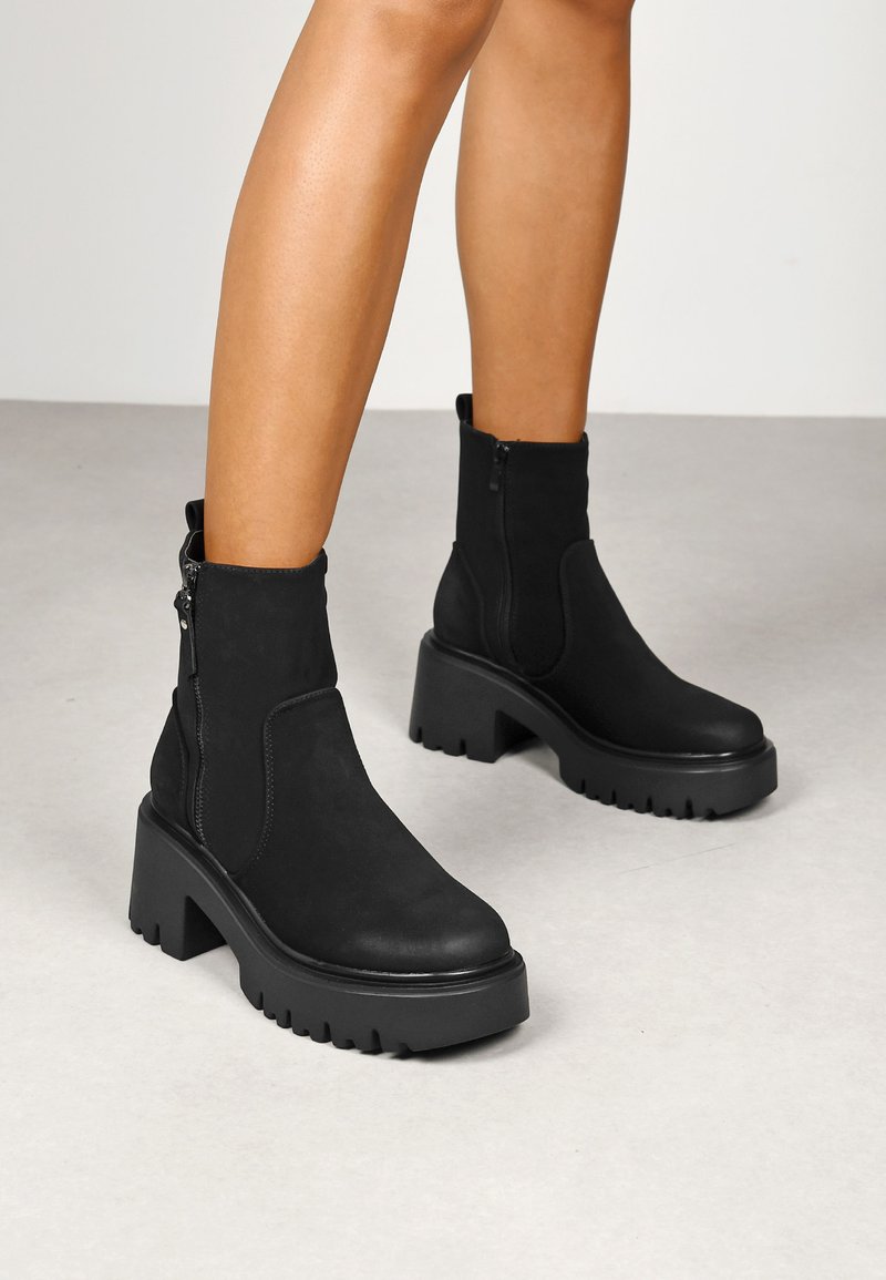 Botines negros de material similar al ante, con suela de plataforma gruesa, cremalleras laterales y un acabado liso y mate.