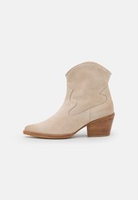 Béžová semišová kotníková bota s špičatou špičkou, zakřiveným horním okrajem a naskládaným dřevěným podpatkem. Obsahuje šití podél boků.