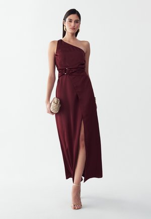 Frau trägt ein ärmelloses bordeauxrotes Kleid mit Oberschenkelschlitz, hält eine gewebte Clutch und trägt goldene Creolen und High Heels.