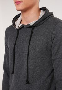 Felpa grigia con una consistenza morbida, caratterizzata da un rivestimento interno grigio chiaro e cordini neri. Il design include un cappuccio regolabile.
