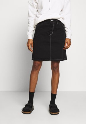 Sorte denimnederdel med hvid kontrastsyning, knæ-længde design, frontlukning med knapper, parret med sorte sokker og sandaler.