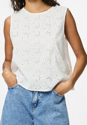 Vrouw draagt een witte mouwloze eyelet kanten top en lichtblauwe hoogtailles spijkerbroek met handen in de zakken, tegen een witte achtergrond.