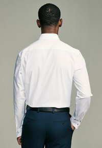 Weißes Hemd mit langen Ärmeln, glattes Material. Tragen mit dunkelblauer maßgeschneiderter Hose, ergänzt durch einen strukturierten schwarzen Gürtel.