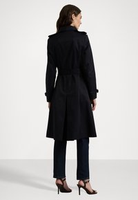 Lange zwarte trenchcoat met een brede kraag, ceintuur in de taille en een achteropening, gecombineerd met donkere jeans en sandalen met banden. Gladde stofstructuur.