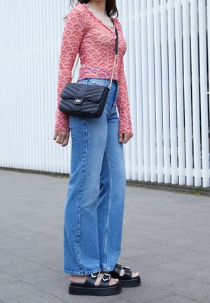 Femme portant un haut à manches longues à motifs roses, un jean bleu à jambes larges, des sandales plateformes noires, et un sac bandoulière noir matelassé.