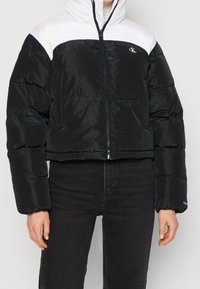 Černobílé bundy do písku s kapucí, vpředu na zip, s logem CK na prsou, nošeno s černými kalhotami.