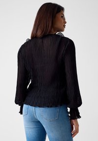 Czarna plisowana bluzka z długimi rękawami, przezroczystą fakturą, falbankowymi mankietami i zaokrąglonym dołem. Połączona z niebieskimi dżinsami, podkreślającymi szczupłą sylwetkę.