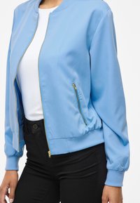 Hele sinine bomber-jakk, mis on valmistatud siledast kangast, esitades esikäepideme, soonikkoes krae ja ühe lukuga küljetasku. Koos mustade pükstega.