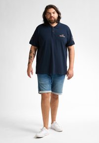 Marinblå polo med korta ärmar, treknapplåsning och diskret logotyp. Kombinerad med uppvikta jeansshorts och vita sneakers.