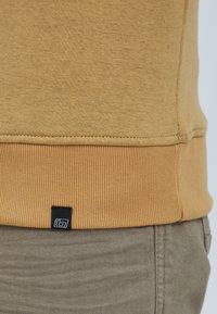 Bluza koloru musztardowej żółci z prążkowanym wykończeniem na dole i mankietach, z małą czarną metką u dołu. Miękka, teksturowana tkanina.