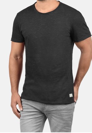 Uomo che indossa una maglietta grigia scura a maniche corte con collo rotondo e jeans grigi chiari, in piedi con le mani rilassate lungo i fianchi.