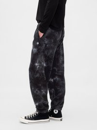 Άτομο φορώντας μαύρο και γκρι tie-dye παντελόνι φόρμας με χέρια στις τσέπες, μαύρα ψηλά αθλητικά παπούτσια και μαύρο μακρυμάνικο μπλουζάκι, στέκεται στο πλάι.