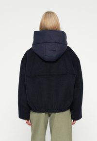 Persona rubia que lleva una chaqueta acolchada negra con una gran capucha y pantalones verde claro, de pie de espaldas contra un fondo blanco.