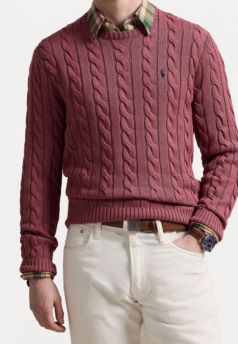 Homme portant un pull torsadé bordeaux par-dessus une chemise à carreaux verte, un pantalon blanc, une ceinture marron et une montre-bracelet avec un cadran bleu foncé.