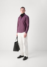 Langsleeve, overhem met knopen in dieppaars, gecombineerd met witte broeken. Zwarte leren schoenen en een zwarte tote bag maken de look compleet.