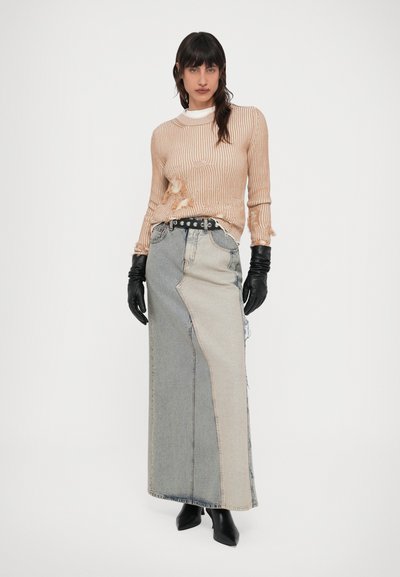 Femeie purtând un pulover bej tricotat uzat, fustă lungă din denim în două nuanțe, mănuși negre din piele, curea cu ținte și cizme negre cu vârf ascuțit.