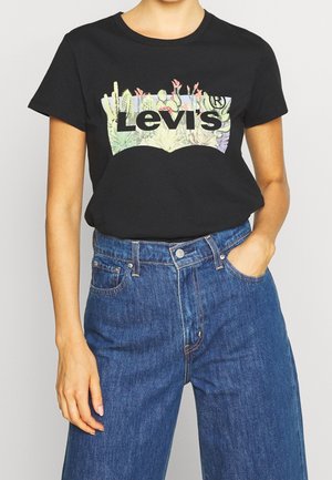 Osoba nosząca czarny t-shirt Levi's z kwiatowym logo i niebieskie jeansy z wysokim stanem, jedna ręka w kieszeni, przycięte na wysokości klatki piersiowej i ud.