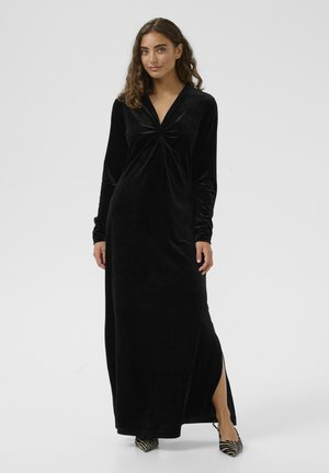 Langes schwarzes Samtkleid mit V-Ausschnitt, gerafften Details an der Brust, langen Ärmeln und einem seitlichen Schlitz. Kombiniert mit gemusterten High Heels.