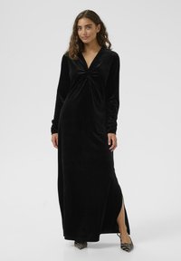 Lång svart sammetklänning med V-ringning, samlad detaljer vid bysten, långa ärmar och en sidoklyvning. Matchad med mönstrade högklackade skor.