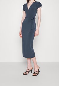 Donkerblauwe omslagjurk met korte mouwen, een diepe V-hals en een strikdetail in de taille, gecombineerd met zwarte, open sandaaltjes met hoge hakken en bandjes.